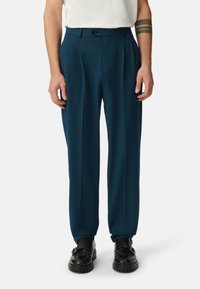 Gepaste broek in diep teal, gemaakt van zachte stof. Heeft een rechte pijp, plooien aan de voorkant en een enkele knoopsluiting. Zwarte schoenen.