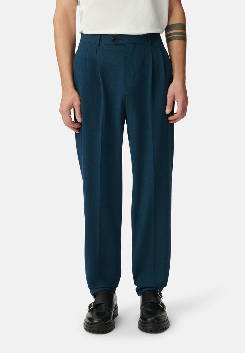 Gepaste broek in diep teal, gemaakt van zachte stof. Heeft een rechte pijp, plooien aan de voorkant en een enkele knoopsluiting. Zwarte schoenen.