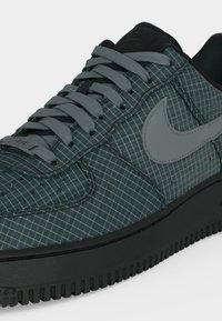 Σκούρο πράσινο αθλητικό παπούτσι Nike με γκρι μοτίβο πλέγματος, γκρι κορδόνια και γκρι λογότυπο Nike σε μαύρη σόλα.