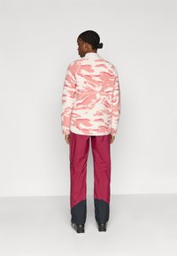 Fleecejakke med pink og hvidt camouflage-design, kombineret med vinrøde vandtætte bukser med sorte detaljer ved kanten.
