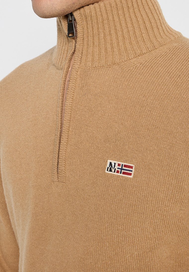 Tan gebreide trui met een hoge kraag, halve rits en een klein geborduurd logo op de borst. Zachte textuur en geribbelde halsontwerp.