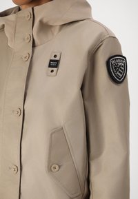 Giacca beige con cappuccio, chiusura a bottoni e un patch sul maniche sinistro. Realizzata in tessuto liscio con un leggero lucido e una forma strutturata.