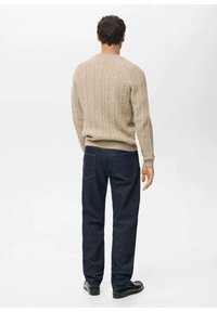 Maglione beige lavorato a maglia con motivo a treccia, abbinato a jeans blu scuro. L'outfit presenta polsini e orlo a coste, con scarpe nere.