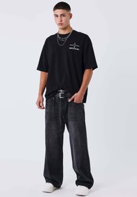 Camiseta negra oversize con logo gráfico, combinada con pantalones vaqueros anchos de mezclilla negra descolorida y zapatillas blancas. Presenta collares de plata en capas.
