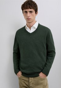 Suéter verde de lana con cuello en v, puños y dobladillo acanalados, usado sobre una camisa blanca abotonada, combinado con pantalones beige. Textura suave.
