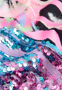 Mehrfarbiger Stoff mit großen pinken und schwarzen Drucken, entlang eines Randes mit blauen und violetten Pailletten verziert, die eine strukturierte und glänzende Optik erzeugen.