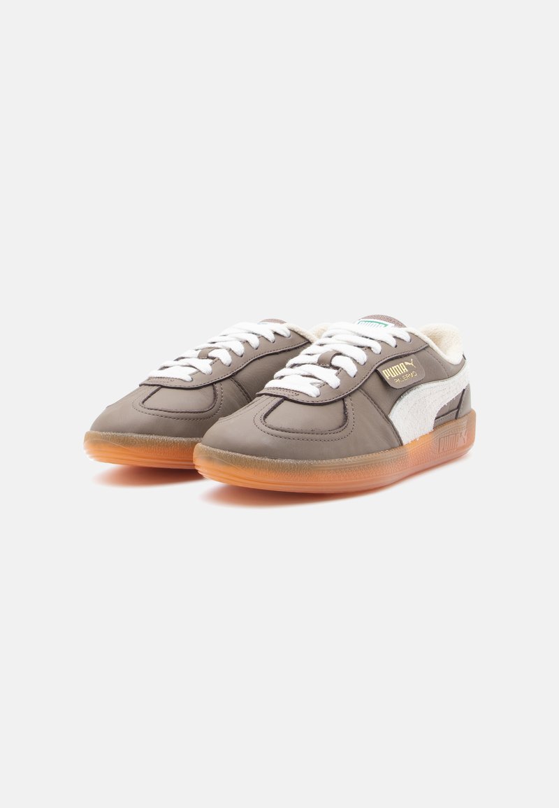 Par de zapatillas Puma grises con cordones blancos, franja lateral blanca y suelas naranjas translúcidas sobre un fondo liso.