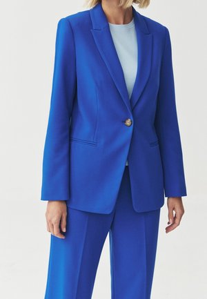 Person iført en knaldblå skræddersyet blazer med en enkelt knap over matchende blå bukser og en lyseblå top.