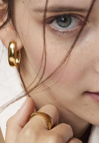 Złota kolczyk w kształcie obręczy o gładkiej, błyszczącej powierzchni oraz cienki złoty pierścionek z teksturowanymi akcentami noszony na palcu, z widoczną skórą na zdjęciu.