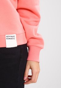 Sudadera coral con puños de canalé, con una etiqueta blanca que dice "NUEVA OLA ROMÁNTICA". Llevada sobre unos pantalones vaqueros oscuros, mostrando una textura suave.