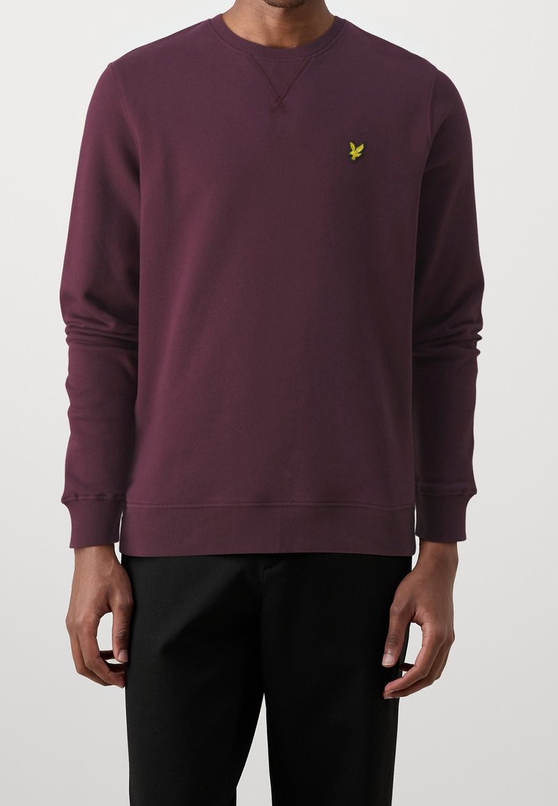 Homme portant un sweat-shirt à manches longues violet foncé avec un petit logo jaune d'oiseau sur la poitrine gauche et un pantalon noir.