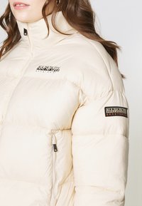 Lichtbeige pufferjack met een hoge kraag, voorzien van een logo op de borst en een geborduurd patch op de mouw. Ritszakdetails.