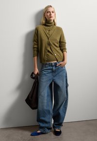 Cardigan a maglia verde oliva con colletto alto e bottoni, abbinato a jeans blu larghi e scarpe basse blu scuro, mentre tiene una borsa a tracolla scura.