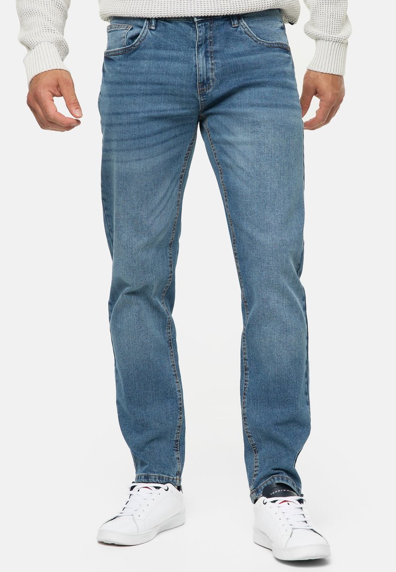 Homme portant un jean bleu droit, des baskets blanches et un pull en maille de couleur claire, debout devant un fond uni.