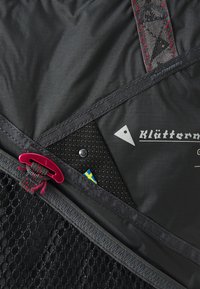 Svart stoff ryggsekk detaljer med meshlomme, røde hardware aksenter og en logo med en trekant. Sysømmer med kontrasterende mønstre.