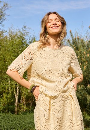 Lachende vrouw in beige kanten jurk met cirkelpatronen staat buiten met handen in de zakken en gesloten ogen in fel zonlicht.