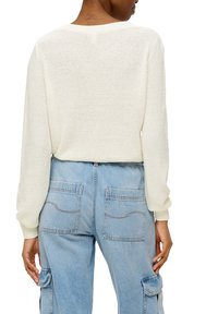 Weißer, strukturierter Pullover mit langen Ärmeln, kombiniert mit hellblauen Cargo-Hosen, die über Fronttaschen und sichtbare Nähte verfügen.