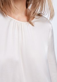 Blusa di un leggero beige con tessuto morbido, scollo rotondo e pieghe sottili sul fronte, caratterizzata da maniche lunghe e una vestibilità rilassata.