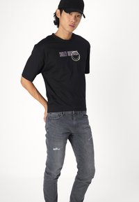 Camiseta de algodón negra con el gráfico "SMILEY ORIGINALS" en púrpura y amarillo. Combinada con unos vaqueros grises desgastados. Ajuste casual y relajado.