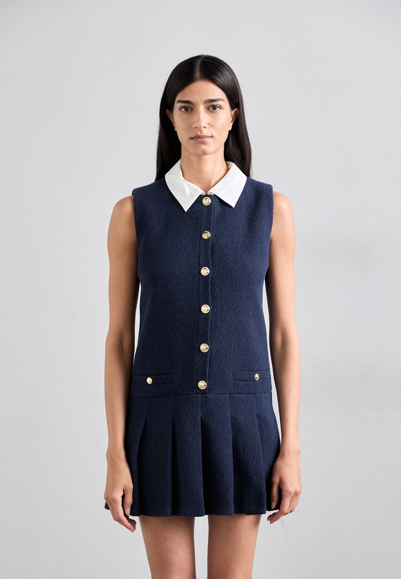 Robe sans manches bleu marine avec un tissu texturé, un col blanc et des boutons dorés sur le devant. Elle dispose de deux poches avant et d'une jupe plissée.
