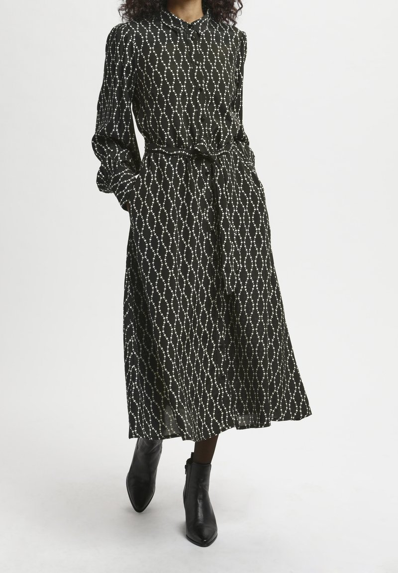 Femme portant une robe midi noire à manches longues avec un motif chaîne blanche, taille ceinturée et bottines noires, debout devant un fond uni.