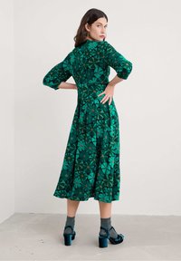 Žena pózuje s rukama v bocích, na sobě má zelené květinové midi šaty, šedé ponožky a tyrkysové saténové sandály na podpatku, na jednoduchém pozadí.