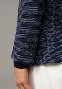 Marineblaue strukturierte Blazerärmel mit einem dezenten geometrischen Muster, ausgestattet mit vier schwarzen Knöpfen und einem marineblauen gestrickten Bündchen.
