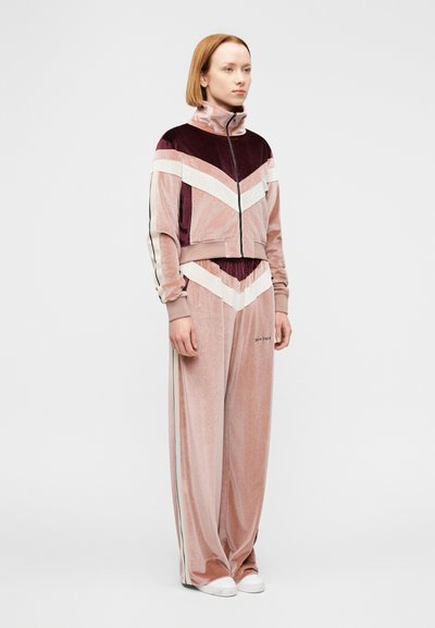 Αθλητικό βελούδινο σετ σε αποχρώσεις του ροζ, του μπορντό και του εκρού. Κοντό μπουφάν με φερμουάρ και σχέδιο σε σχήμα βέλους· φαρδιά, ψηλόμεση παντελόνια.