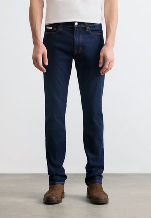 Mann trägt dunkelblaue Slim-Fit-Jeans und braune Stiefel, steht auf grauem Boden vor weißem Hintergrund.