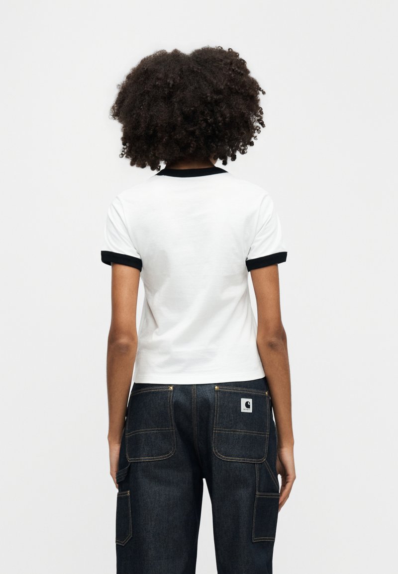 Maglietta bianca a maniche corte con bordi neri, dal design aderente. Abbinata a pantaloni in denim scuro, con tasche visibili e cuciture evidenti.