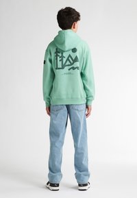 Felpa verde menta con stampa grafica sul retro, abbinata a jeans azzurri chiari e scarpe da ginnastica nere. Vestibilità rilassata con cappuccio regolabile.