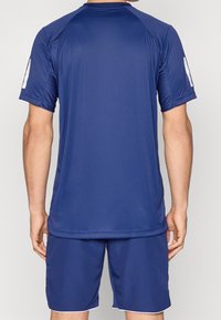 Osoba w jednolitym, granatowym, krótkim rękawie koszulce sportowej oraz dopasowanych szortach, widoczna z tyłu na neutralnym tle.