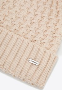 Cappello beige lavorato a maglia con motivo strutturato, polsino a coste e targhetta metallica con marchio. Materiale morbido e caldo, ideale per il clima freddo.