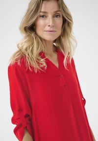 Röd knapp-up skjorta med texturerad tyg, med V-hals, rullade ärmar och subtila linjer, presenterad mot en ljus bakgrund.