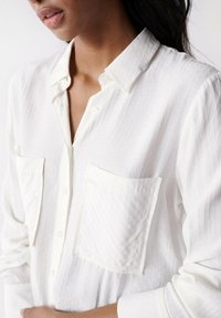 Chemise blanche à manches longues avec rayures texturées, deux poches avant et un col boutonné. Tissu doux et léger avec une coupe décontractée.