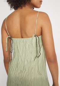 Απαλό πράσινο, υφασμάτινο φόρεμα με λεπτές τιράντες, διακοσμημένο με ρυθμιζόμενους κόμπους στην πλάτη, δημιουργώντας ένα ρυτινωτό εφέ.