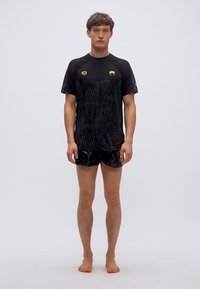 Μαύρο αθλητικό μπλουζάκι με υφασματικό σχέδιο, κίτρινα λογότυπα και μαύρο σορτς με διακριτικό μοτίβο. Το μοντέλο στέκεται ξυπόλυτο.