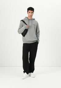 Felpa grigia con cappuccio e tasca frontale, borsa a tracolla nera, pantaloni larghi neri e sneakers bianche, su uno sfondo semplice.