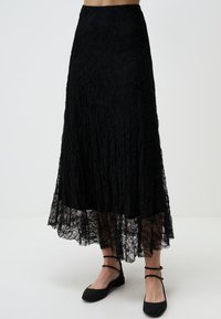 Jupe maxi en dentelle noire avec un design à volants, dotée d'un ourlet festonné et de motifs floraux délicats, assortie de sandales plates noires.