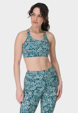 Vrouw draagt een bijpassende blauwgroene sportbeha en legging met bloemenprint, staat ontspannen en kijkt opzij.