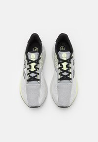 Zapatillas deportivas de color gris claro con cordones negros y parte superior de malla texturizada, con un acento amarillo y una suela acolchada.