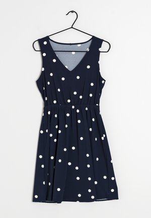 Marineblaues ärmelloses Kleid mit weißen Polka-Dots, V-Ausschnitt, tailliert, an einem schwarzen Kleiderbügel vor weißem Hintergrund hängend.