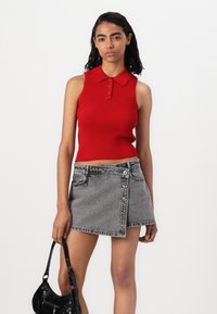 Top polo sans manches rouge côtelé avec trois boutons, associé à une mini-jupe en denim gris avec une coupe asymétrique et des détails de boutons.