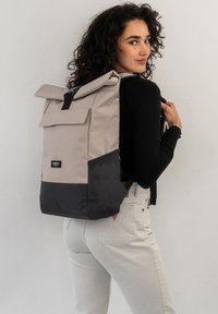 Roll-top backpack in beige and dark gray. Made of durable fabric, featuring a magnetic closure, adjustable straps, and side pockets.
