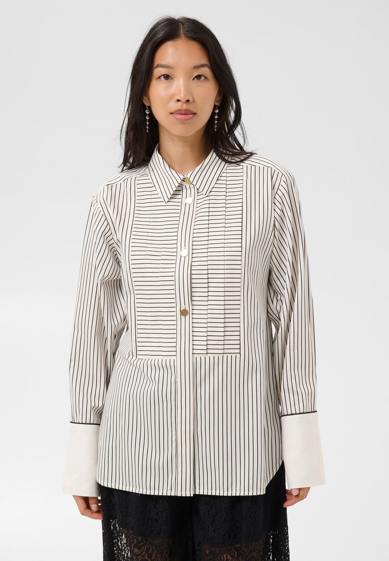 Femme portant une chemise blanche boutonnée avec des rayures verticales noires et de larges poignets blancs, assortie à un pantalon en dentelle noir.