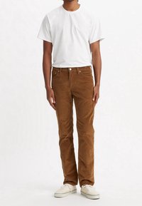 Biała koszulka z okrągłym dekoltem, połączona z brązowymi sztruksowymi spodniami. Spodnie mają przednie kieszenie i zapięcie na guzik, a do tego noszone są tenisówki.