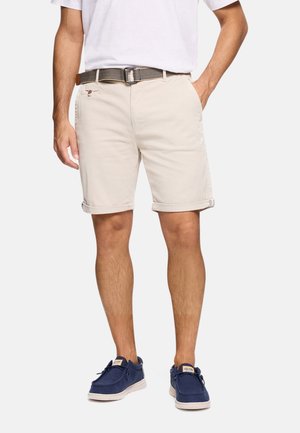 Hombre con pantalones cortos beige con dobladillo enrollado y cinturón beige y negro, camiseta blanca y zapatos casuales azul marino, de pie con una mano en el bolsillo.