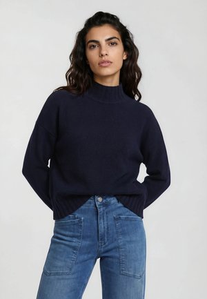 Woman wearing a dark navy ribbed turtleneck sweater and high-waisted blue jeans with large front pockets, standing against a plain background.