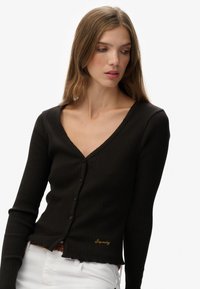 Mujer con cabello largo castaño que lleva un cárdigan negro de canalé con botones y pantalones blancos, mirando hacia abajo y de lado contra un fondo blanco.