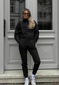Completo sportivo nero con maniche lunghe, colletto alto e fascia elastica in vita. Sneakers bianche con dettagli neri e calzini bianchi corti.